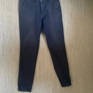 Mörkblå chinos med tapered fit - Snygga mörkblå chinos med tapered passform och klassiska fickor fram och bak. Byxorna har knapp och dragkedja i midjan samt bälteshällor. Perfekta för en clean och stilren look, passar dig som gillar enkel design och bekväma material. Storlek 33 32