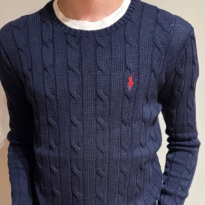 Marinblå stickad tröja från Polo Ralph Lauren - Snygg marinblå stickad tröja från Polo Ralph Lauren med klassiskt kabelstickat mönster och röd broderad logga på bröstet. Rund halsringning och långa ärmar. Perfekt för dig som gillar stilrena och tidlösa plagg. Storleken är M(junior) men passar xs 