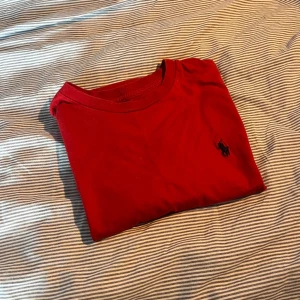 Röd t-shirt från Polo Ralph Lauren - Riktigt snygg röd t-shirt från Polo Ralph Lauren med klassisk svart broderad logga på bröstet. T-shirten är i mjuk bomull och har en enkel, stilren design med rund hals och korta ärmar. Perfekt för en clean och avslappnad look.