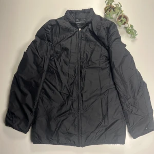 Svart jacka Peak Performance  - Svart pufferjacka från Peak Performance, modell Melfort J. Jackan har en stilren look med hög krage och dragkedja framtill. Tillverkad i 100% polyamid, perfekt för kyligare dagar. Klassisk passform och långärmad design.