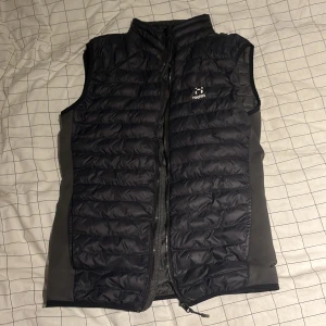 Svart dunväst från Haglöfs, storlek S - Svart dunväst från Haglöfs med quiltad design och medel hög krage. Västen har dragkedja framtill och praktiska fickor. Materialet är polyester och dun, perfekt för lager-på-lager under kyliga dagar. Diskret Haglöfs-logga på bröstet och insida med extra fickor