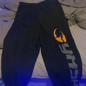 Svarta loose träningsbyxor från Gasp - Svarta träningsbyxor från GASP i storlek L med bred resår i midjan. Byxorna har en loose passform och ett stort tryck i orange och grått längs ena benet. Perfekta för chill eller sport, tillverkade i mjukt syntetmaterial.