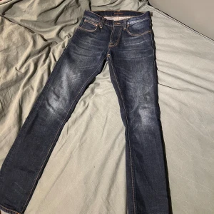 Mörkblå jeans från Nudie Jeansco W28 L32 - Snygga mörkblå jeans från Nudie Jeansco med klassisk femficksdesign och orange kontrastsömmar. Modellen har raka ben och knappgylf. Jeansen är tillverkade i slitstarkt bomullsmaterial och har en stilren look med diskret logga på bakfickan. Byxorna sitter som 28 i midjan 