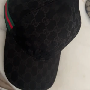 Svart Gucci keps med GG-mönster - Snygg svart keps från Gucci med klassiskt GG-monogram över hela kepsen. Har en grön och röd rand på sidan som ger den typiska Gucci-looken, kommer med tag o dustpåse