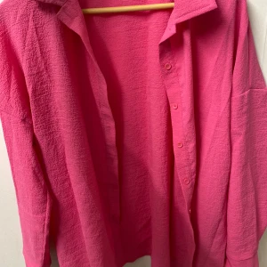 Oversized rosa skjorta med struktur - Säljer en oversized skjorta i starkt rosa färg med krage och knappar framtill. Skjortan har lång ärm och är tillverkad i ett strukturerat