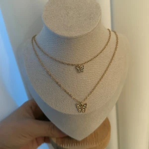 Guldigt halsband med fjärilar - Dubbelkedjat halsband i guld med två små fjärilshängen dekorerade med glittriga stenar. Kedjorna är tunna och eleganta, och fjärilarna ger en söt och trendig touch. Perfekt för dig som gillar detaljer och vill sticka ut lite extra.