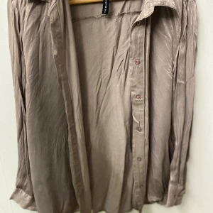 Beige silkeslen skjorta  - Snygg beige skjorta  med silkeslen känsla och klassisk krage. Skjortan har långa ärmar och knappar framtill, perfekt för en stilren look. Materialet ger ett lyxigt intryck och passar dig som gillar en avslappnad men ändå dressad stil.