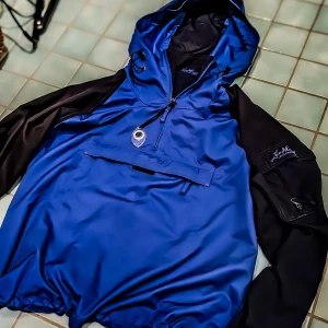 Blå vindjacka från Lundhags - Säljer en blå och svart vindjacka från Lundhags med huva och snörning. Jackan har en stor ficka framtill med lock, half zip och är vattentät. Perfekt för dig som gillar outdoor och vill ha en snygg jacka med grym passform.