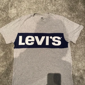 Grå Levi's t-shirt med blå logga - Snygg grå t-shirt från Levi's i 100% bomull. Framsidan har en stor mörkblå panel med vit Levi's-logga som ger en cool kontrast. Klassisk rund hals och korta ärmar, perfekt för en chill och stilren look.