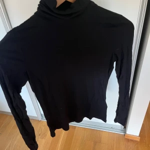 Svart polotröja i bomull - En svart, klassisk polotröja med lång ärm och hög krage. Tröjan är i mjuk bomull och har en slim passform som sitter snyggt på kroppen. Perfekt att styla med jeans eller under en jacka för en clean look.