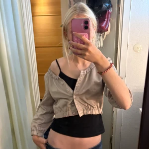 Beige bolero från Zoul - Säljer en beige croppad blus från Zoul med trekvartsärm och rundad hals. Blusen har knappar framtill och elastisk kant vid midjan och ärmslut. Perfekt att styla med linne under för en chill look.