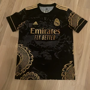 Real Madrid x Adidas svart/guld tröja S - Snygg Real Madrid fotbollströja från Adidas i svart med guldiga detaljer och drakmönster. Tröjan har korta ärmar, klubbmärke och Adidas-logga på bröstet samt 'Emirates Fly Better' tryck. Tillverkad i lätt och ventilerande AEROREADY-material.