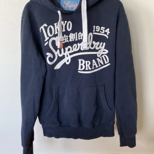 Mörkblå Superdry hoodie med tryck - Mörkblå hoodie från Superdry med stort vitt tryck på bröstet och coola japanska tecken. Tröjan har huva med vita snören och en stor magficka. Perfekt för dig som gillar streetstyle och vill ha något bekvämt och snyggt.