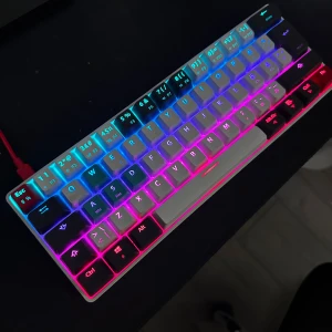 NOS C450 Mini Keyboard Pro RGB - Kompakt mekaniskt tangentbord med RGB-belysning och nordisk layout. Perfekt för gaming och skrivbord med begränsat utrymme. Tangenterna är i gott skick, vissa har utbytta färger. USB-anslutning. Modell: C450 Mini Keyboard Pro RGB.