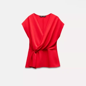 Röd omlottopp från Zara - Snygg röd omlottopp från Zara med v-ringning och knyt i sidan. Toppen är ärmlös och har en lätt figursydd passform som framhäver midjan. Perfekt att styla med jeans eller kjol för en trendig look.