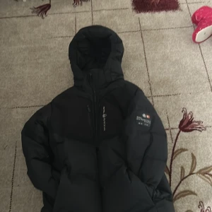 Möts i handen Sthlm  - Säljer en svart pufferjack  med luva  och dragkedja framtill. Jackan har en patch med logga på armen och är riktigt varm och skön för kalla dagar. Perfekt för dig som vill ha en stilren och funktionell vinterjacka.❤️