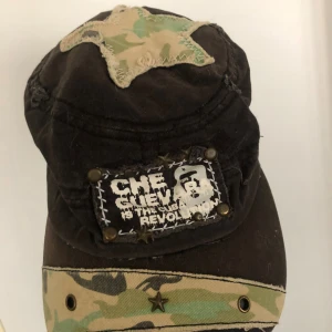 Svart keps med Che Guevara och stjärnor - Unik svart keps med Che Guevara-tryck framtill, dekorerad med nitar och stjärnor. Har en stor tygstjärna i camouflage på ovansidan och ett camouflagemönstrat band med metallstjärna på skärmen. Kepsen är tillverkad i bomull och har en cool, rebellisk vibe.