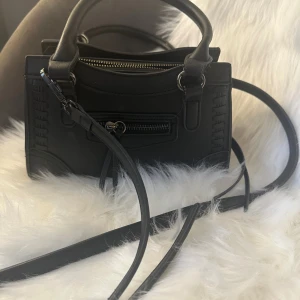 Svart handväska med dragkedja - Snygg svart handväska i skinnimitation med både handtag och lång axelrem. Väskan har en cool detalj med dragkedja framtill och dekorativa sömmar på sidorna. Perfekt storlek för det viktigaste och passar till många olika outfits.
