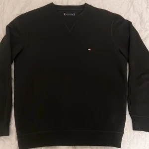 Svart sweatshirt från Tommy Hilfiger - Svart sweatshirt från Tommy Hilfiger med liten broderad logga på bröstet. Tröjan har rund hals, ribbade muddar och lång ärm. Materialet är mjuk bomull med inslag av polyester, perfekt för chill dagar. Enkel och stilren design som funkar till jeans eller joggers. Jag säljer den för att den inte passar min klädsel och den är bara använd nån gång. Den passar även M då den är lite större i storleken. Ny pris är 1200