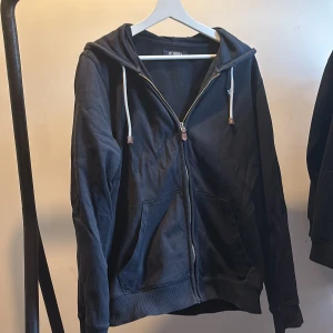 Svart hoodie från Morris med dragkedja - Svart hoodie från Morris med dragkedja, huva och vita snören. Broderad logga på bröstet och två stora fickor framtill. Skön och enkel modell i bomull, perfekt för chill dagar eller när du vill ha en stilren look.