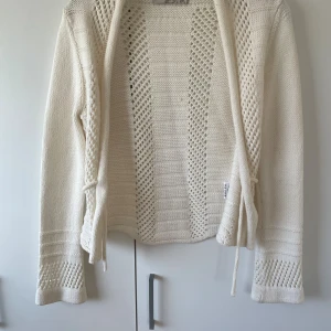 Vit stickad kofta  - Säljer en vit stickad kofta från CLARA i storlek M. Koftan har snygga virkade detaljer, knytband framtill och långärmade ärmar med mönstrad struktur. Materialet är en mix av bomull och akryl, vilket gör den både mjuk och luftig.