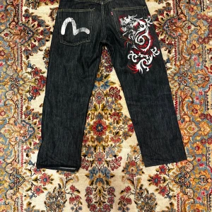 Evisu jeans med drakemönster bak - Snygga mörkblå evisu jeans med rak passform och coolt drakemönster i vitt och rött på bakfickan. Klassisk femficksmodell med kontrastsömmar och en unik detalj på bakfickan. Perfekta för dig som vill sticka ut lite extra.