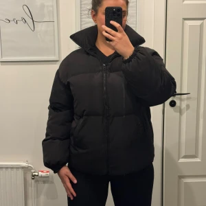 Svart pufferjacka med hög krage - Säljer en svart pufferjacka med hög krage och dragkedja framtill. Jackan har en loose passform och är tillverkad i syntetmaterial. Perfekt för kalla dagar och har en stilren look som funkar till många outfits.