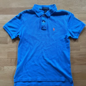Blå pikétröja från Polo Ralph Lauren - Klassisk blå pikétröja från Polo Ralph Lauren med orange broderad logga på bröstet. Tröjan har korta ärmar, krage och knappar framtill. Tillverkad i bomull för en skön och luftig känsla.