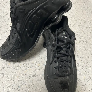 Svarta Nike Shox sneakers strl 41 - Nike Shox sneakers i helsvart med ikoniska fjädrande Shox-sulan. Snygg sportig design med rund tå, snörning och diskret Swoosh-logga på sidan. Ovandel i syntet och mesh för bra ventilation. Perfekt för dig som gillar retrovibe och streetstyle.