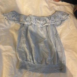 Ljusblå offshoulder blus med broderi - Supersöt ljusblå offshoulder blus med broderade blommor och håldetaljer längs kanten. Blusen har smockad nederkant och är perfekt för varma dagar. Luftig och lätt i materialet, med romantisk vibe.