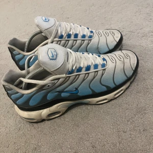 Nike Air Max Tn blå och vita sneakers - Nike Air Max Tn sneakers i en fräsch mix av vitt, blått och grått med svarta detaljer. Snyggt vågigt mönster på ovandelen och klassisk Air Max-sula. Skorna har snörning och rund tå, tillverkade i syntetmaterial och mesh för skön komfort.