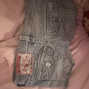 True Religion ljusblå jeansshorts - Säljer ett par snygga ljusblå jeansshorts från True Religion med ikoniska bakfickor och grova kontrastsömmar. Shortsen har råa kanter, klassisk femficksdesign och silverfärgade knappar. 