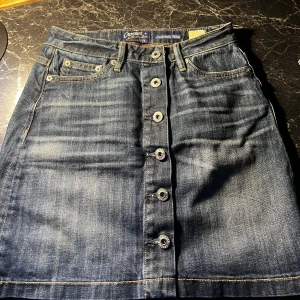 Jeanskjol med knappar från Crocker - Säljer en snygg jeanskjol från Crocker med klassisk blå tvätt och kontrastsömmar. Kjolen har fem knappar framtill, fickor både fram och bak samt hällor för bälte. Perfekt för dig som gillar en enkel och cool denimstil.