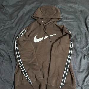 Brun Nike hoodie med logga och huva - Säljer en brun hoodie från Nike med stor vit swoosh-logga på bröstet och svarta band med vita loggor längs ärmarna. Tröjan har huva med snörning och är långärmad. Perfekt för dig som gillar sportig och avslappnad stil. Aldriga använd