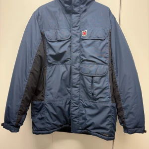 Blå parkajacka från Fjällräven - Parkajacka från Fjällräven i mörkblå och svarta toner, med flera fickor framtill och dragkedja. Jackan har lång ärm, hög krage och slitstarka detaljer. Materialet är syntet och polyester, perfekt för kalla dagar. Klassisk Fjällräven-logga på bröstet. Dragkedjan på bilden saknar en tand, men går att dra upp och ner ändå. 