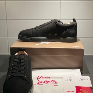 Svarta mocka sneakers från Christian Louboutin - Snygga svarta sneakers från Christian Louboutin i mjuk mocka med klassisk röd sula och diskreta metalldetaljer på sidan. Skorna har rund tå, platt sula och snörning framtill. Perfekt för dig som vill ha en lyxig och stilren look med en ikonisk touch helt nya inte alls använd. 