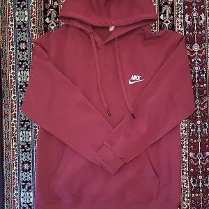 Vinröd Nike hoodie med broderad logo - Säljer en vinröd hoodie från Nike med broderad vit logga på bröstet. Tröjan har huva med snörning, känguruficka och långa ärmar. Perfekt för chill dagar eller när du vill ha en sportig look.