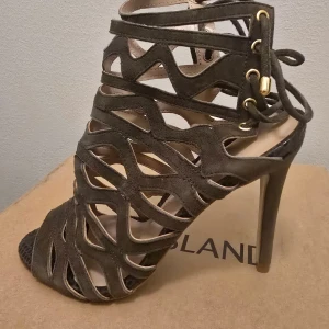 Khakigröna klackade sandaler River Island - Säljer ett par khakigröna sandaler från River Island med höga klackar och öppen tå. Skorna har ett coolt utskuret mönster i mocka, snörning med gulddetaljer och plattformsula baktill. Perfekta för dig som vill sticka ut med en edgy stil.