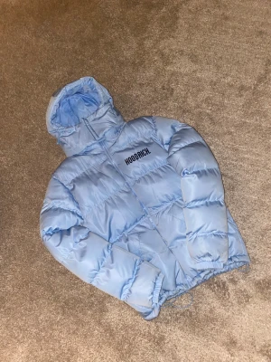 Ljusblå pufferjacka från Hoodrich - Säljer en ljusblå pufferjacka från Hoodrich i storlek S. Jackan har huva, dragkedja framtill och stora Hoodrich-loggor både på bröstet och ryggen. Materialet är glansigt och syntetiskt, med quiltad design och elastiska muddar vid ärmslut och nederkant.