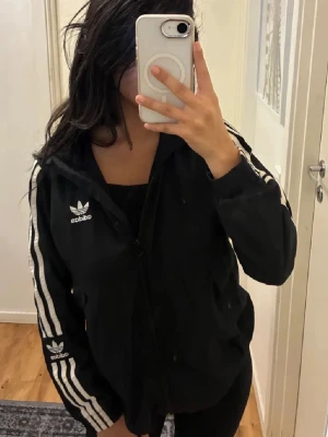 Svart Adidas vindjacka med vita ränder - Svart vindjacka från Adidas med klassiska vita ränder längs ärmarna och broderad logga på bröstet och ärmen. Jackan har huva, dragkedja framtill och två fickor. Perfekt för en sportig och chill stil. Materialet är lätt och syntetiskt.