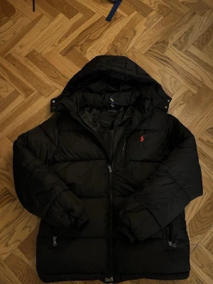 Svart dunjacka från Polo Ralph Lauren - Svart dunjacka från Polo Ralph Lauren med huva och röd broderad logga på bröstet. Jackan har dragkedja framtill, flera fickor med dragkedja och quiltad design. Perfekt för kalla dagar och har en clean, stilren look. Måtten stämmer dock inte! 🙌🏽