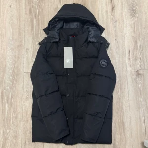 Svart pufferjacka från Canada Goose - Svart pufferjacka från Canada Goose med stor huva och broderad logotyp på ärmen. Jackan har knappar och dragkedja framtill, quiltad design och är långärmad. Perfekt för kalla dagar med sin rejäla dunfyllning och stilrena look.