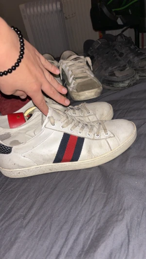 Gucci sneakers med röd och blå rand - Säljer ett par vita Gucci sneakers med klassiska röd- och blåa ränder på sidan. Skorna har snörning, röd detalj med stjärna på hälen och svart mönstrad hälkappa. Tillverkade i skinn med textildetaljer. Perfekta för dig som vill sticka ut med en ikonisk look.