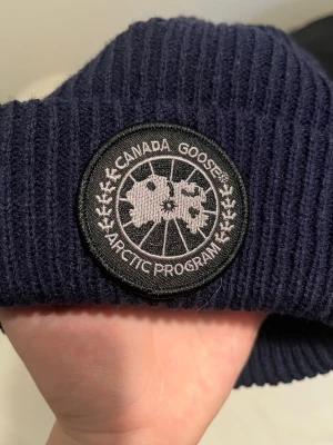 Mörkblå ribbstickad mössa Canada Goose - Snygg mörkblå ribbstickad mössa från Canada Goose med klassisk rund logotyp-patch framtill. Mössan har uppvikt kant och är tillverkad i mjukt stickat material som håller dig varm under kalla dagar.