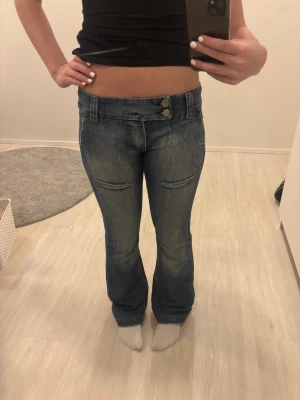 Blå bootcut jeans från Just Jeans - Snygga blå bootcut jeans från Just Jeans med låg midja och coola slitningar. Jeansen har dubbla knappar fram, kontrastsömmar i gult och unika bakfickor med lock och knapp. Perfekt passform och klassisk jeanslook med lite tvättad effekt.