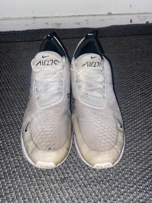 Nike Air Max 270 vita sneakers - Nike Air Max 270 sneakers i vitt med svarta detaljer och stor synlig Air-enhet i hälen. Ovandel i mesh och syntet, snörning framtill och rund tå. Sulan är genomskinlig med gul ton undertill. Perfekta för dig som gillar sportig och modern stil.