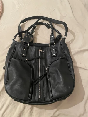 Perfekta skolväskan❤️ Rymlig - Snygg svart axelväska i skinn med flera praktiska fack och silverfärgade detaljer. Väskan har två stora ytterfickor med dragkedja, samt en mittficka med dragkedja och rejäla handtag. Perfekt för dig som vill ha plats för allt och ändå se cool ut. Lite sliten på handtagen och längst upp syns på två sista bilderna. Inget som syns mycket eller stör funktionen