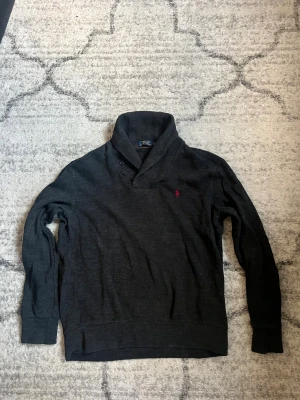 Mörkgrå kofta från Polo Ralph Lauren - Snygg mörkgrå kofta från Polo Ralph Lauren med bred krage och två knappar vid halsen. Tröjan har lång ärm och en liten röd broderad logga på bröstet. Perfekt för dig som gillar klassisk stil med en modern twist.