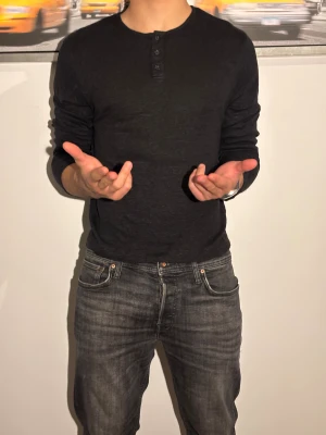 Svart Henley - Svart långärmad tröja med tre knappar vid halsen. Tröjan har en rund halsringning och är tillverkad i ett mjukt och lätt material som känns skönt mot huden. Passformen är normal och stilen är enkel och clean, perfekt för dig som gillar minimalistiska plagg.