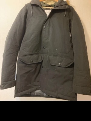 Svart parkajacka från Jack & Jones - Parkajacka i svart från Jack & Jones med stor huva och beige fuskpäls. Jackan har två stora fickor med knappar framtill, dragkedja och tryckknappar. Insidan är quiltad och fodrad i grönt och grått syntetmaterial. Perfekt för kalla dagar. ⚠️Köparen betalar för frakt⚠️